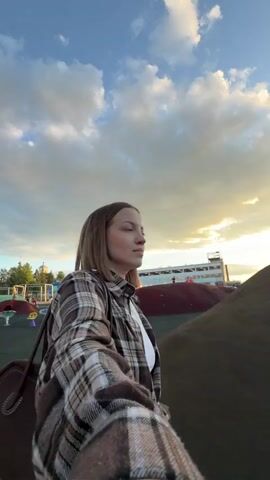 A woman in a plaid shirt is walking up a mound on a playground as the camera records in selfie mode. The sky is a mix of blue and cloudy with the setting sun in the background. A playground is visible behind the mound. A man appears carrying a young child, and both are descending the mound.