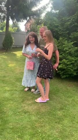 The video shows three people in a grassy outdoor setting, likely a backyard. On the left is a woman in a light blue floral dress, holding a pink gift bag and a glass of white liquid. She is smiling and looking towards her right. In the center is a man wearing a green t-shirt with the word "LACOSTE" on it, and jeans. He is holding a glass of white liquid and looking towards the woman on the right. On the right is a young woman in a black floral dress and purple slippers. She is touching her hair and looking towards the man. In front of the people is a large arrangement of colorful flowers. The background shows a wooden fence, trees, and a house in the distance. The sun is shining, casting shadows on the grass. The overall mood appears to be casual and celebratory.