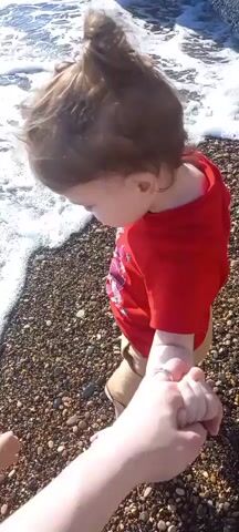 This is a close-up, over-the-shoulder shot of a toddler being led by the hand along a pebble beach. The toddler is wearing a bright red t-shirt with a graphic design and tan shorts, and has their hair pulled up in a ponytail. The toddler's left hand is held by an adult, whose hand is visible in the foreground. The sea is to the left of the frame, with gentle waves lapping at the shore, creating white foam. The sun is shining brightly, casting a warm glow on the scene. The overall mood is one of carefree exploration and parental guidance. The location cues and time of day suggest a pleasant, sunny day at the beach, likely in the morning or afternoon.
