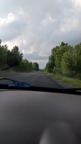 The video offers a first-person perspective from inside a moving car, looking out through the front windshield. The dashboard and a portion of the windshield wipers are visible at the bottom of the frame. The vehicle is traveling on a straight, two-lane asphalt road. Lush green trees line both sides of the road, creating a dense, verdant canopy overhead. The sky is overcast with a mix of white and grey clouds. In the distance, another car can be seen approaching on the same road. At approximately 0:03, a triangular warning road sign with a red border becomes visible on the right side of the road. The setting appears to be a rural or forested area, consistent with the provided location context of Ablyazovo, Russia. No specific people are visible, nor is any discernible text present in the scene. The activity depicted is driving.