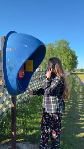 A medium shot captures a young woman with long, straight brown hair standing outside on a sunny day, engaged in a phone conversation at a blue public telephone booth. She is dressed in a plaid shirt and patterned pajamas, holding a red phone to her right ear and resting her left hand near the phone's keypad. Her face expresses a mix of surprise and amusement. The phone booth is blue with a red interior, displaying some writing and a yellow note. In the background, a white wooden fence runs parallel to a dirt path, with green grass and trees visible under a clear blue sky.