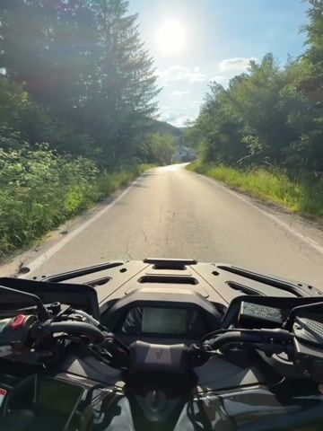 This is a first-person perspective video recorded from the handlebars of a dark-colored ATV. The ATV is traveling down a paved, two-lane road that winds through a rural, forested area. The road surface is somewhat worn with visible cracks and a white line marking the edge.

The ATV's dashboard is visible in the foreground, featuring a digital display screen. The handlebars are black with various controls, including a red grip on the left.

The setting is a sun-drenched countryside with lush green trees lining both sides of the road. Tall pine trees are particularly prominent on the left. In the distance, a white house with a red roof is visible nestled amongst the trees on a slight incline. The sun is bright and high in the sky, casting a strong glare and illuminating the scene. The sky is blue with some scattered white clouds.

The time of day appears to be late morning or early afternoon, given the sun's position. The weather seems to be clear and sunny. The overall mood conveyed is one of peaceful exploration and enjoyment of a scenic drive in nature. There is no visible text in the video. The location context provided is Boltata, Bulgaria.