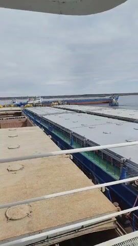 The video captures a maritime scene featuring multiple large barges being maneuvered on a body of water, likely a river or canal in Rybinsk, Russia. The shot is taken from an elevated position, possibly from another vessel or a dock, with railings visible in the foreground.

In the mid-ground, at least three long, flat-topped barges are visible, linked together in a line. Their tops are covered, suggesting they are carrying cargo. The barges have blue hulls, and the gaps between them show the water. At the far end of this convoy of barges, a blue and white tugboat is positioned, appearing to either push or pull the barges along the waterway. Further in the background, another larger vessel, possibly a cargo ship or another tug, can be seen on the horizon. A few crew members are faintly visible on the far barge and the tugboat, overseeing the operation.

The activity taking place is the transportation or maneuvering of these barges. The tugboat is actively engaged in controlling the movement of the barge convoy. The water is calm, indicating placid conditions. The sky is overcast or partly cloudy, suggesting a daytime scene, possibly mid-morning or afternoon, with ample ambient light but no direct sunlight. No discernible text is visible on any of the vessels or in the surrounding environment. The overall impression is one of typical commercial waterway operations.