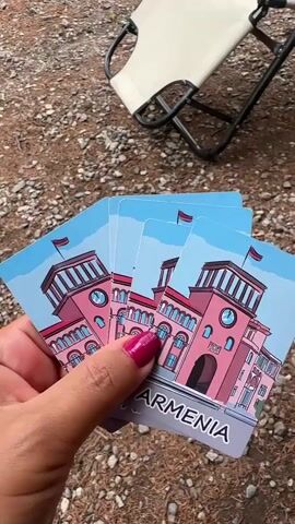 The video shows a close-up of a person's hand holding playing cards against a backdrop of gravel and a portable chair. In the first few seconds, the hand holds playing cards with clover symbols and numbers. Then, the cards are replaced by cards featuring illustrations of a pink building with a clock tower and the word "ARMENIA" written on them. The person's fingernails are painted a vibrant pink with black polka dots. The setting appears to be outdoors, possibly a campsite or picnic area, given the gravel ground and the presence of the portable chair. The lighting suggests it might be daytime. There is no explicit indication of the time of day or weather conditions, beyond the natural light. The activity involves displaying or revealing cards, which could be part of a game or a presentation of collectible items. The focus on the cards and the hand holding them suggests a personal or perhaps instructional context.