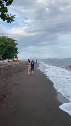 Two people walk along a dark sand beach, the ocean waves lapping at the shore to their right. The sky is cloudy and the sun is low. In the distance, other people can be seen walking along the beach and swimming in the water. A few boats are pulled up on the sand to the left, and trees with green leaves overhang the beach. The video is shot from a slight angle, looking out towards the ocean.