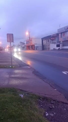 The camera is positioned on a sidewalk overlooking a wet roadway, with a patch of green grass at the bottom of the frame. A traffic sign stands on the left side of the sidewalk. Several buildings line the right side of the road, some of which are businesses with signs in Spanish. The sky is overcast, and it appears to be early morning or late evening, with streetlights illuminated and car headlights visible in the distance. A light-colored car drives by on the road, heading towards the camera.
