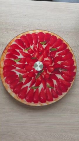 This video shows a close-up, top-down view of a round object with a strawberry tart design on a wooden surface. The object has a golden-brown crust around the edges, a white filling, and is covered in sliced red strawberries arranged in a circular pattern. There are also a few green mint leaves scattered among the strawberries. In the center of the object is a silver, circular knob or handle, suggesting it might be part of a clock or a lazy Susan. The camera slowly zooms out, revealing more of the wooden table surface. The lighting is even, and the focus remains sharp on the object throughout the video.