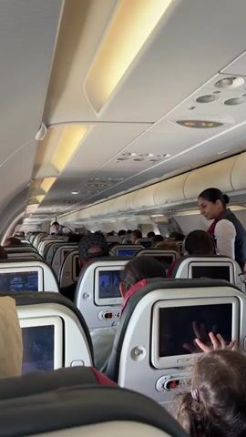 A flight attendant in a dark vest over a red shirt walks down the aisle of a crowded airplane. She bends over to interact with a passenger. Rows of seats with individual screens are visible on either side of the aisle. Several passengers are seated, some with headrests up. The overhead compartments and lighting fixtures are visible along the ceiling of the plane.