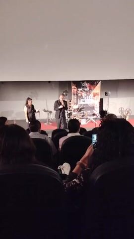 The video shows an indoor scene of an auditorium-style movie theater, filled with seated viewers. The focus is on a stage area where a man, dressed in black, is speaking into a microphone, gesturing with his hands as he addresses the audience. To his left stands a woman in a dark dress. Behind them is a banner, possibly promoting a film festival, with the word “CINE” prominently displayed. In the foreground, individuals in the audience can be seen, some holding up cell phones, possibly recording the event. The overall atmosphere appears to be that of a presentation or introduction before a film screening.