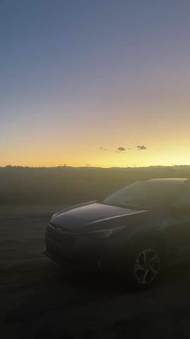 The video shows a dark-colored Subaru parked on a dirt road at sunrise. The car is facing towards the left of the frame. The sky is a gradient of colors, transitioning from blue at the top to yellow and orange near the horizon. There are a few small clouds in the sky. The landscape in the background appears to be a field or hills. The overall scene is dimly lit, creating a silhouette effect on the car.