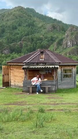 A person sits on a bench in front of a wooden cabin playing a guitar. The cabin has a corrugated metal roof and is surrounded by green grass and trees. In the background, a large, forested mountain rises under a cloudy sky. The camera is positioned at a medium distance from the cabin.