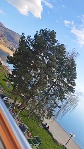 The video is a slightly tilted landscape view of a serene lake on a sunny day with a partly cloudy sky.  The foreground includes a patch of green grass, several trees, and a sidewalk along the shoreline. There are a few people sitting on the shore in the distance.  A metal railing is visible at the bottom of the frame.  The lake extends to the horizon, where distant mountains can be seen. The water appears calm with reflections of the sky. The overall atmosphere is peaceful.