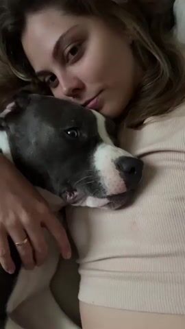 A close-up video shows a woman embracing a pit bull dog. The woman's face is partially visible as she rests her head against the dog's. The dog's head is turned towards the camera, with its eyes open and its mouth slightly ajar. Both the woman and the dog appear to be in a relaxed position, possibly lying down. The background is blurred, suggesting an indoor setting. The lighting is soft, illuminating the faces of the woman and the dog.