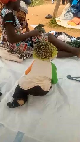 A video shows a person, likely a child, sitting on a white cloth spread on the ground. The person is wearing a white shirt with orange and green accents on the shoulders and collar, black pants, and dark socks. A yellow net covers their head. A woman in a patterned dress and headscarf is seated to the left of the child, with clothing piled around her. Behind them, there are more piles of clothing spread out on a white tarp, and several other people are visible in the background. The ground appears to be dirt. A hand reaches into the frame from the right and briefly touches the child.