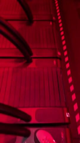 The media file shows a dimly lit fitness studio with predominantly red lighting. The scene is filled with rows of treadmills and exercise benches, indicating a gym setting. Several people are present, engaged in various fitness activities. In the foreground, a woman is visible on a treadmill, seemingly mid-workout. Further back, other individuals can be seen on treadmills and around the benches, suggesting a class or workout session is in progress. The overall atmosphere appears energetic and focused, typical of a group fitness environment. The red lighting creates a dramatic and intense mood. No text is visible in the media file. The location is identified as Palo Alto, United States of America. The time of day is not explicitly indicated but the indoor setting and lighting suggest it could be any time.