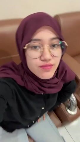A young woman with a maroon hijab, black top, and gold-rimmed eyeglasses is seen in a close-up selfie video. She has pink lipstick and visible braces, which she shows while smiling. She adjusts her hijab and playfully looks at the camera from different angles, exhibiting a cheerful demeanor. The background shows a brown sofa or chair and a light-colored wall with a decorative, possibly stucco, pattern. The setting appears to be indoors in Kaliwates, Indonesia.