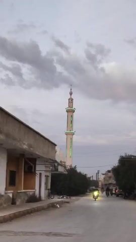 The video shows a street scene with a mosque in the background. The sky is cloudy and it appears to be dusk. A white mosque minaret, decorated with green lights, stands tall against the sky. The street is lined with buildings, some of which are houses. Several motorcycles with their headlights on are driving down the street. The audio captures the Islamic call to prayer.