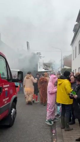 The scene unfolds on a street where a parade is taking place, amidst a cloudy and overcast atmosphere. A red firetruck labeled "FEUERWEHR 112" (Fire Department 112) is visible on the left side of the frame, partially obstructing the view. The focus is drawn to a large, white structure resembling an airplane or float, emitting thick white smoke, suggesting it's part of the parade. Numerous people are lining the street, many dressed in costumes. Notable costumes include individuals in lion, bear, and pink bunny pajamas, adding a whimsical element to the event. Some people are interacting with each other, and the general atmosphere appears festive. The environment suggests a small town setting, with buildings visible in the background, although obscured by the smoke and overcast sky.