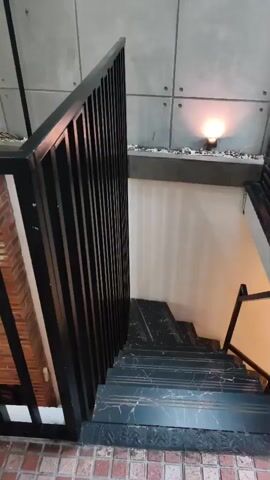 The video shows a modern interior with a black metal railing and a dark marble staircase. The staircase leads down to a lower level. The walls are made of large concrete panels with exposed screws. There are small spotlights on the wall, illuminating decorative stones. Further into the space, a wooden deck is visible, furnished with a low black coffee table with a glass top and a gray sofa. A large window offers a view of the exterior. A slatted black metal screen is positioned to the left, separating the stairwell from the seating area. A potted plant is visible near the screen. The lighting is warm and ambient.
