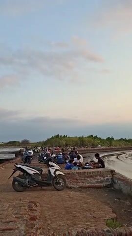 The video shows a serene coastal scene at what appears to be dusk or dawn. The sky is painted with soft, pastel colors from the setting or rising sun, with scattered clouds creating a dynamic backdrop. A body of water, possibly a river or sea, stretches across the frame, reflecting the sky's gentle hues.

In the foreground, a weathered, brick-lined pathway or pier runs along the water's edge. The path is unpaved and appears somewhat rugged. Several motorbikes are parked along this path, indicating a public gathering or a popular spot. One white and black motorbike is prominently featured in the foreground, while others are visible further down the path.

Further along the path, groups of people are seen sitting or standing, some appearing to be engaged in conversation or enjoying the view. Their presence suggests a communal gathering, perhaps for a sunset viewing or a local event. Beyond the water, a lush green landscape with trees forms a distant horizon, contrasting with the more muted tones of the foreground and sky. The overall atmosphere is peaceful and tranquil, suggesting a moment of shared appreciation for the natural beauty of the location.