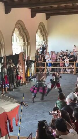 A reenactment battle is occurring in a large hall with arched windows and wooden ceiling beams. A crowd of spectators is gathered behind a wooden barrier, watching two men in medieval armor duel with swords. One duelist wears a dark leather cuirass and a kilt-like garment with a pink and purple plaid pattern, while the other wears a white tunic with red trim and a black vest adorned with white stars. A man in a yellow and brown tunic stands to the side, appearing to officiate the duel. On the left side of the arena, several other men in armor stand ready. The floor is made of dark stone tiles. In the foreground, the backs of spectators are visible, some holding up phones to record the event. The overall atmosphere suggests a historical reenactment or jousting event.