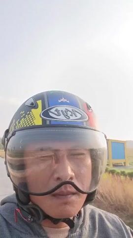 A close-up shot shows a person wearing a black and yellow motorcycle helmet with a clear visor. The person's face is visible through the visor, and they appear to be blinking. The person is wearing a gray hooded jacket. The background is a blur of green and yellow fields and a bright, hazy sky, suggesting an outdoor setting, possibly during daytime. The camera is moving, indicating the person is likely on a moving vehicle, such as a motorcycle. The timestamp indicates the video is very short, just over 5 seconds long.