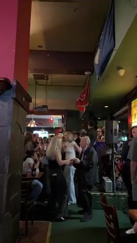 The video shows a group of people dancing and enjoying themselves in what appears to be a crowded bar or pub with a live band playing in the background. There are a few tables and chairs around the room where some people are sitting and drinking. Flags hang from the ceiling. One elderly man dances with two women near the middle of the room. The atmosphere appears lively and festive.