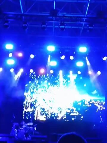 The video shows a view of a concert stage from the audience. Above the stage are several spotlights illuminating the scene in a vibrant blue hue. A large screen behind the musicians displays an animated image, initially a swirling pattern of light, which then transitions into a depiction of a destroyed cityscape. A few audience members are visible in the foreground, their heads blocking a small portion of the lower frame. The camera remains mostly stationary, capturing the dynamic lights and animations on the stage.