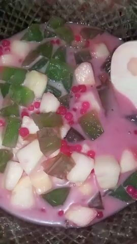 The video shows a close-up shot of a bowl containing a dessert with a pink liquid base. Inside the bowl are cubes of what appears to be coconut meat, green jelly or gelatin cut into rectangular shapes, and small, bright red spherical tapioca pearls. A white spoon is seen stirring the ingredients in the bowl. The bowl is placed inside what appears to be a metal strainer or rack. No people are visible.