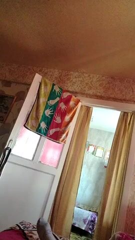 The video shows an interior view of a room, possibly a bedroom or living area. A brightly colored towel with handprints is draped over an open white door or partition. To the right of the door, yellow curtains hang, partially obscuring another doorway leading to a different area of the room. This area appears to have a bed with patterned bedding and possibly framed pictures on the wall. The camera then pans to the left, revealing a mirrored wardrobe and a shelf unit with various items on it, including what looks like a stuffed toy. Further to the left, a television is mounted above a cabinet. The overall impression is of a somewhat cluttered but lived-in space. The video is shaky and appears to be filmed from a low angle, possibly from the perspective of someone lying down.