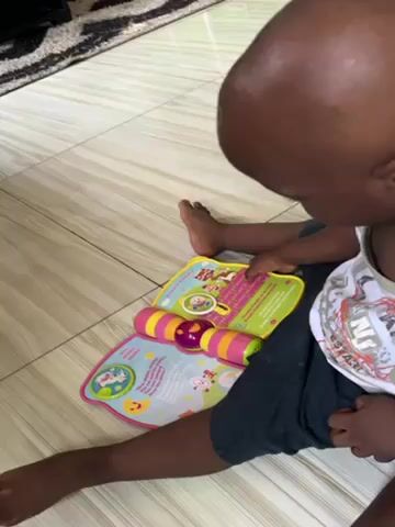 A small, dark-skinned child sits on a light-colored, tile floor with a colorful interactive book open on their lap. The child is wearing a white tank top with a colorful pattern and dark shorts. Their feet are bare. The book is open to a page with pictures and text. The book has a brightly colored, textured roller on the left side. The child is pointing at the book with their right hand. In the background, a small part of a dark-colored rug with a light-colored pattern is visible. The scene is well-lit.
