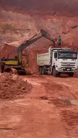 This video shows an excavator loading dirt into the bed of a white dump truck at a construction site. The ground is made of red dirt. A person stands on top of the dirt pile to the right of the dump truck, appearing to be supervising or directing the operation. The excavator is yellow and appears to be older. The dump truck is white and has a large, segmented bed. The excavator's arm is extended, and its bucket is full of dirt, in the process of dumping it into the truck's bed. The environment is an open pit mine or a large excavation site with steep dirt walls. The lighting suggests it is daytime.