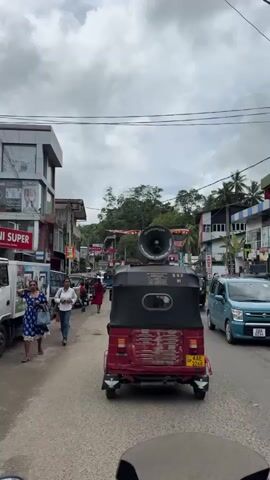 A street scene in Sri Lanka. A red and black tuk-tuk with a large speaker on top is driving down a paved road. Other vehicles, including more tuk-tuks and motorcycles, are also visible. There are buildings on either side of the road, some with shops. People are walking on the sidewalks. The sky is cloudy.