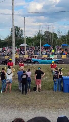 The video shows a demolition derby event. In the foreground, a group of spectators stands behind a chain-link fence, watching the event. Several people in the spectator area are wearing casual clothing. Behind the fence, near the demolition derby track, several individuals in safety gear, including yellow hard hats and red vests, are present. They appear to be officials or safety personnel. Two cars are on the track: one is a blue car that appears to have been damaged and is positioned at an angle, and another dark-colored car is partially visible to the left, also showing signs of damage. In the background, a large crowd of spectators is seated in bleachers, watching the event. The sky is cloudy, and the scene suggests an outdoor event during daylight hours.