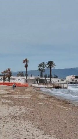 The video shows a beach on a windy day with waves crashing on the shore. A building stands on the beach, partly on stilts over the water. In the background, mountains stretch across the horizon under a cloudy sky. A person is walking along the beach near a construction barrier. The beach itself is covered with a mix of sand and seaweed.