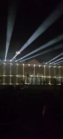 The video shows an outdoor nighttime event. A building facade is illuminated with a light projection that creates a geometric pattern and a central figure. Multiple spotlights from above cast beams of light into the sky. In front of the facade, silhouetted figures are visible, suggesting a performance or audience. The foreground is dark, with the shapes of people visible in silhouette.