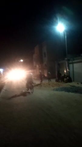 The video presents a dark, nighttime street scene, filmed vertically. A prominent, bright street light is visible on the far left of the frame, casting a strong, white glow. The foreground and middle ground consist of a dark road surface, possibly asphalt or dirt.

In the lower-center of the frame, intense, bright headlights from a vehicle (likely a motorcycle or a car) create a significant glare and lens flare, obscuring details directly behind them.

Several dark, silhouetted figures, presumably people, are gathered in the mid-ground on the right side of the road, near what appears to be a building or wall. Some figures seem to be sitting or squatting, while others are standing.

Around the 0:01 mark, a dark, four-legged animal, possibly a dog with its tail curled upwards, quickly runs from the right side of the frame towards the left, crossing the road in the mid-ground. Immediately following the animal, another dark figure, appearing to be a person, moves across the road in the same direction.

Towards the end of the clip, a bicycle with its own light can be seen passing, its light blending with the larger vehicle's headlights. The background shows dark silhouettes of buildings against a slightly lighter, dark sky. The overall lighting is low, with strong contrasts from the artificial light sources. No text is visible.