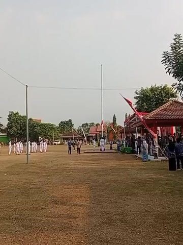 This is a wide shot of an outdoor gathering. A group of people in white uniforms stands in formation on the left side of a grassy field. In the center of the field, a flagpole stands tall, with a red and white flag flying at its top. Several individuals, some in uniform and some in civilian clothes, are gathered around the base of the flagpole. To the right of the flagpole, there is a covered structure with a red roof, decorated with red and white banners. More people are gathered under and around this structure. The sky is overcast. The overall scene suggests an official ceremony or event taking place on school grounds or a similar public space.
