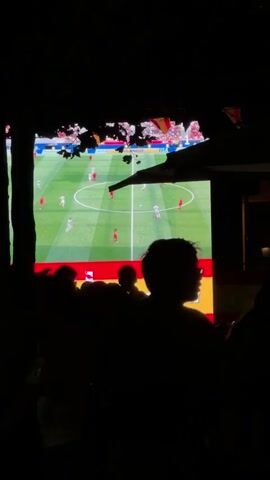 The video shows a crowd of silhouetted people watching a large screen displaying a soccer match. The screen shows players in white and red uniforms on a green field with a white center circle and red and yellow banners at the bottom. The ambient lighting is dark, with the screen being the primary light source. The scene appears to be outdoors or in a dimly lit venue.