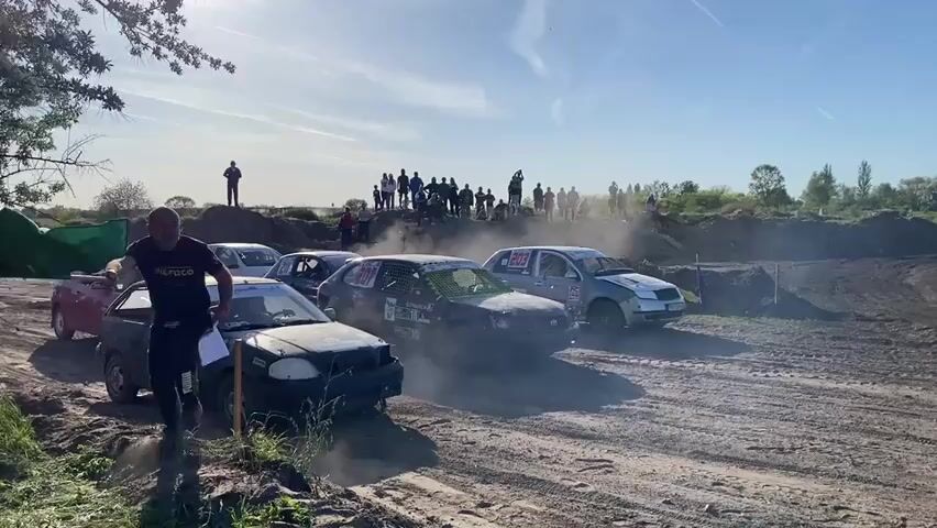 The video shows a group of about five beat-up cars lined up at the start of a dirt track race. Spectators stand behind a small dirt hill in the background, watching. A man with a green flag stands to the left of the cars, waiting to start the race. Dust fills the air as the cars rev their engines. The day is sunny, with a clear blue sky.