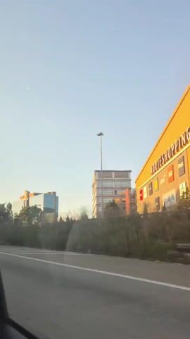 The video shows a view from a moving vehicle, likely a car, looking out a window. The time appears to be late afternoon or early evening, indicated by the warm, golden sunlight illuminating the buildings. The scene features a roadside with some vegetation and debris. In the background, there are several buildings, including a modern glass-fronted building, a taller building with multiple floors, and a long, low building with a yellow facade. Signs on the buildings are visible, including one that says "SPORTSHOP." A construction crane can be seen near one of the buildings on the left. The sky is clear and light blue. The road surface has white lines, suggesting it is a highway or a major road.
