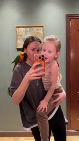 A woman stands holding a young child, presumably her daughter, while taking a selfie in a mirror. A small, colorful parrot is perched on the woman's left shoulder. The woman is wearing a gray T-shirt and black leggings. The child is wearing a tan tank top and brown leggings, with a small red hair tie in her blonde hair. They are in a room with pale green walls and a wooden door. A framed piece of artwork hangs on the wall behind them. The child sticks her tongue out at the camera.