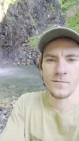 A man with light brown hair and blue eyes, wearing a green cap and a light green t-shirt, takes a selfie video in front of a waterfall. The man is smiling and speaking to the camera. The waterfall is cascading down a rocky cliff face, surrounded by trees. The water is clear and the bottom of the pool is visible. The sun is shining and there is a rainbow visible in the mist of the waterfall.