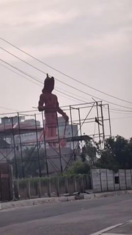The video shows a large, reddish statue of a human-like figure standing behind a metal scaffolding. The statue appears to be incomplete or under construction, as scaffolding is erected around it. The figure is facing to the left and is holding a long object in its right hand, possibly a weapon or staff. The statue has a headdress or crown. Behind the statue, there are buildings and trees. In the foreground, there is a road and a low concrete wall with some fencing. Overhead, power lines are visible against a cloudy sky. The camera is stationary.