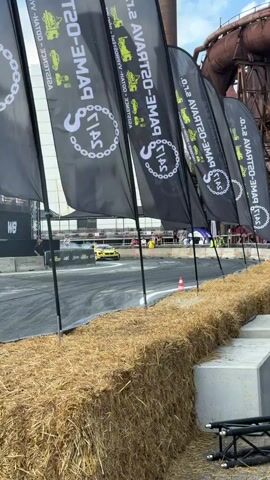 A drift car is performing a maneuver, creating a large plume of smoke. The car is yellow and black with various sponsor logos. It is on a paved track, with hay bales and concrete blocks lining the foreground. In the background, there are large industrial structures and flags with the text "SALT GAME 0-20". There are spectators and staff visible in the distance.