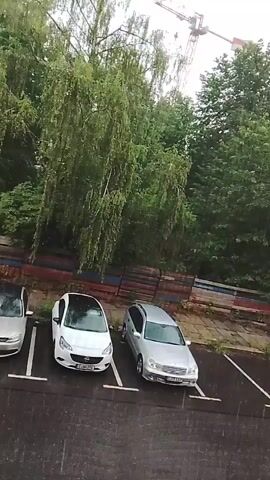 The video shows a view of a parking lot from above. The parking lot is wet, indicating rain. There are several cars parked in designated spots. From left to right, there is a silver car, a white car, a silver station wagon, and a yellow car. In the background, there are trees and a construction crane. The weather appears overcast.