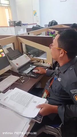The video shows a man wearing glasses and a dark uniform sitting at a desk in an office environment. He is looking at a computer screen and holding a stack of papers. A scanner is visible on the desk next to the computer. The office appears to have cubicle dividers and other desks in the background. The man is holding a mouse in his right hand. The screen shows folders and what appear to be documents. There are papers on the desk and to the left of the computer. The man's uniform has a badge on it.