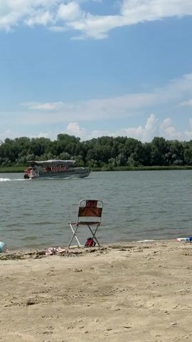 The video shows a tranquil scene on a sandy beach next to a wide river. In the foreground, a weathered camping chair sits on the sand, its fabric showing a reddish-brown pattern. To the left of the chair, a small pile of beach items, possibly a towel and flip-flops, is visible. Further to the right, a child's toy, appearing to be a water gun, rests on the sand. The river occupies the midground, its surface rippled by a gentle breeze. A white boat with a canopy, carrying several passengers, cruises across the water, leaving a white wake behind it. The background is dominated by a dense line of lush green trees that forms the far bank of the river. The sky above is a clear blue, dotted with fluffy white clouds, suggesting a pleasant summer day. The overall impression is one of peaceful relaxation and enjoyment of the outdoors.