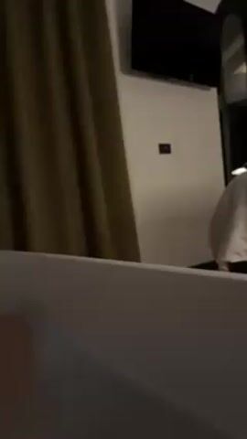 The video shows a view from inside a bathtub.  The tub is white. The camera is positioned at a low angle, looking out into a dimly lit room. A dark green curtain is visible to the left. On the wall, there is a flat-screen television and a small dark switch. To the right, there is a bed with white linens. The room appears to be a hotel room.  No people are clearly visible, though there is a blurry, reddish object visible within the tub.