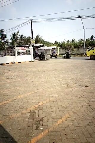 The video shows a street scene with paved ground in the foreground. A yellow truck with a wooden cargo bed is moving to the right. Several motorcycles are also visible on the road. Some are carrying passengers. There are trees and a wall in the background. Power lines are strung across the top of the frame. A few stalls or small structures are present on the left side of the road. The sky is bright and appears to be daytime.